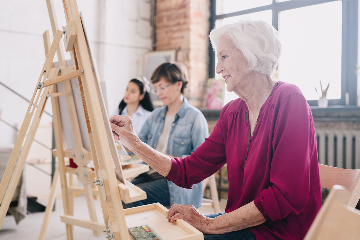 Senior Artist Painting in Art Studio Side view portrait of art students sitting in row and painting at easels in art studio, focus on smiling senior woman enjoying work copy space