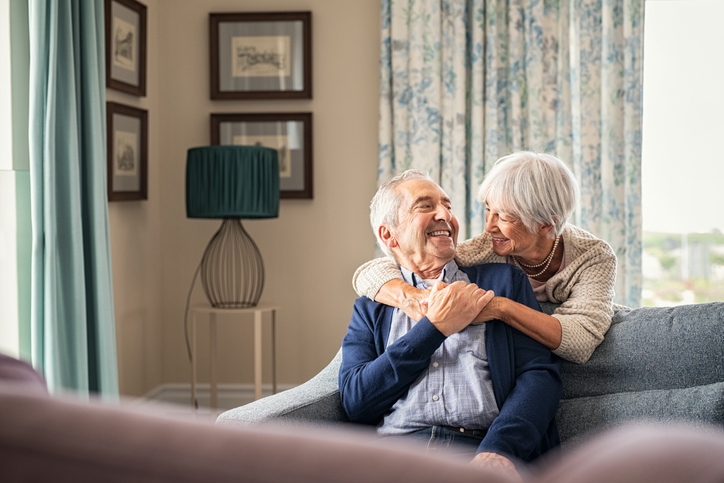 Senior couple hugging and having fun at home Happy senior woman embracing her husband at home while laughing together. Smiling wife hugging old man sitting on couch from behind. Joyful retired couple having fun at home while looking at each other with copy space. Love and unity concept.
