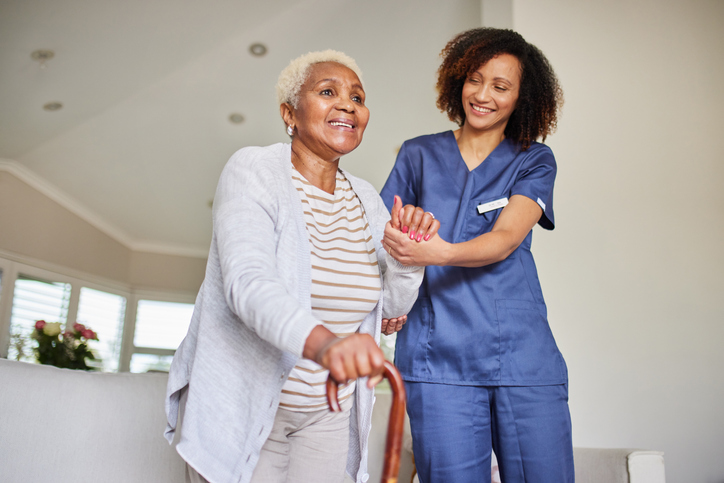 Supporting Steps: Caregiver and Senior Woman Warm scene of a caregiver assisting an elderly woman with a cane. Both are smiling, creating a heartwarming and supportive atmosphere indoors.