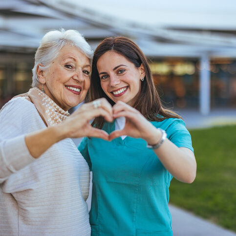 Senior Woman and Caregiver Forming Heart Shape Outdoors A joyful senior woman and her caregiver make a heart shape with their hands, symbolizing love and support in an outdoor setting.